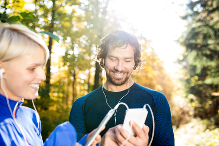 Beautiful couple running together outside in colorful sunny autumn forest using a fitness app on their smartphones. Using phone app for tracking weight loss progress, running goal or summary of their run.の写真素材