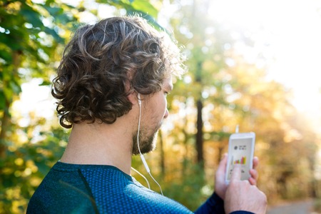 Unrecognizable man running outside in colorful sunny autumn forest using a fitness app on his smartphone. Using phone app for tracking weight loss progress, running goal or summary of his run. Rear view.の写真素材