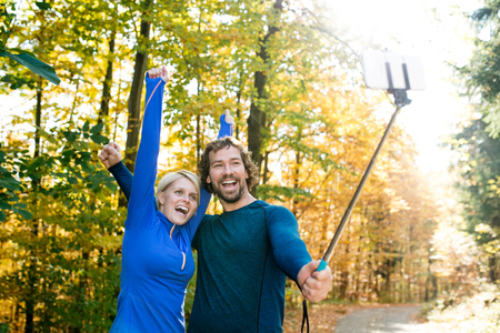 Beautiful young runners in colorful sunny autumn forest taking selfie with smartphone on selfie stickの写真素材