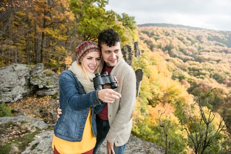 Beautiful young couple on a walk, standing on a rock against colorful autumn forest, woman holding binoculars, pointing at something with her fingerの写真素材