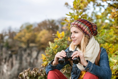 Beautiful young woman holding binoculars against colorful sunny autumn forestの写真素材
