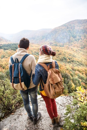Beautiful couple with backpacks standing on a rock against colorful autumn forest, rear viewの写真素材