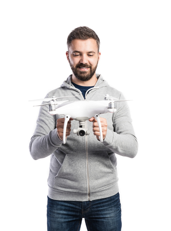 Young hipster man in gray sweatshirt holding drone with camera. Studio shot on white background,...