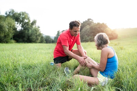 Active senior runners outside in field. Woman with injured knee. Man helping her. Green sunny summer nature.の写真素材