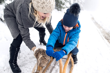 Beautiful young mother pushing her son on sledge, foggy white winter nature.