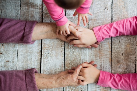 Close up. Unrecognizable grandmother and her granddaughters holding hands. Studio shot on white wooden background.の写真素材