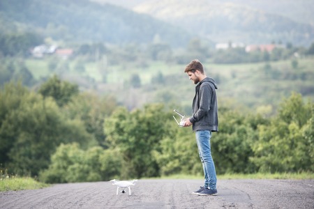Young hipster man with remote control and flying drone with camera. Green sunny nature.