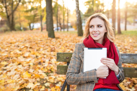 Beautiful young blonde woman in red knitted scarf and checked coat sitting on wooden bench, holding tablet. Sunny autumn park.の写真素材