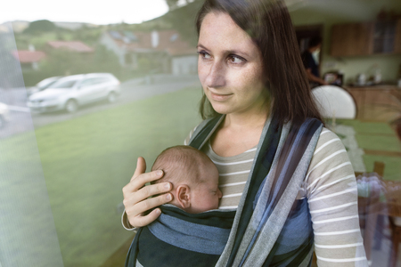 Beautiful young mother with her newborn baby son in sling at homeの写真素材