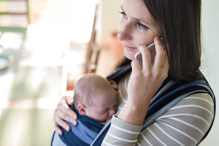 Beautiful young mother with her newborn baby son in sling at home, holding smart phone, making phone...