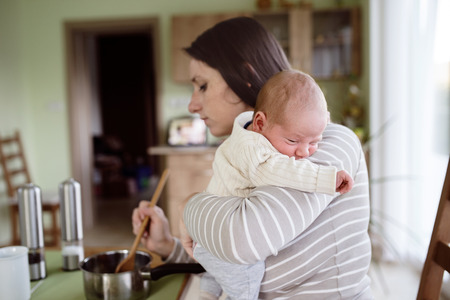 Beautiful young mother at home in the kitchen holding her newborn baby son, cooking, mixing...