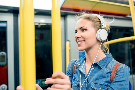 Beautiful young blond woman in denim shirt standing in subway train with white headphones listening musicの写真素材