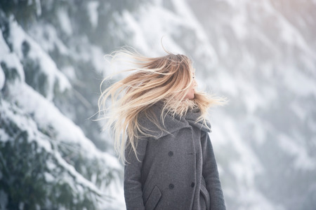 Beautiful young blonde woman in gray coat with fluttering hair covering her face on a walk in winter nature. Snowy day.の写真素材