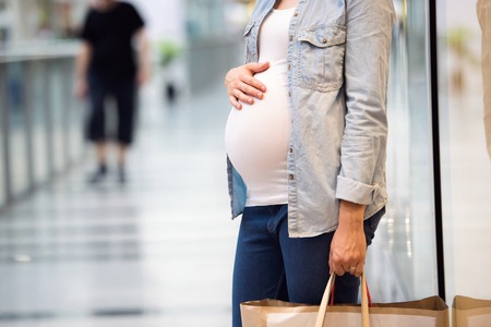 Unrecognizable pregnant woman in white t-shirt and denim shirt touching her belly, holding bags, shoppingの写真素材