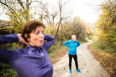 Beautiful active senior couple running together outside in sunny autumn forest, stretching