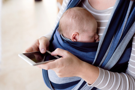 Unrecognizable young mother with her newborn baby son in sling at home, holding smart phone, writing...