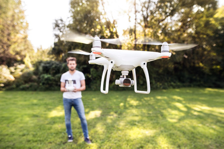 Young hipster man with remote control and flying drone with camera. Green sunny nature.