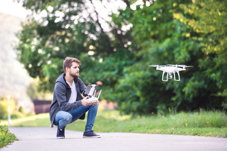 Young hipster man with remote control and flying drone with camera. Green sunny nature.