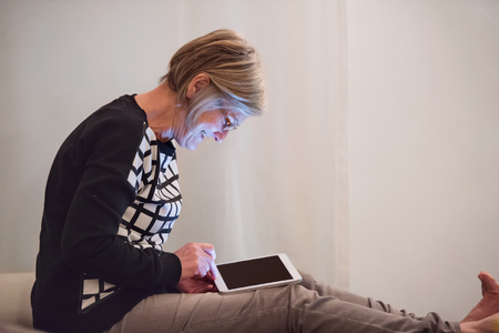 Senior woman with tablet sitting on couch in living room, reading something.の写真素材