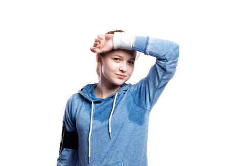 Teenage girl in blue sweatshirt, wearing phone armband, wiping sweat from her forehead. Beautiful young sportswoman, studio shot on white background, isolated.の写真素材
