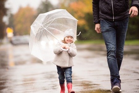 Unrecognizable young father with cute little daughter under the umbrella. Walking in the street on a rainy day.の写真素材