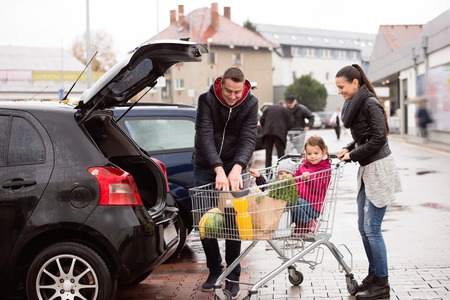 Young parents unloading groceries from shopping cart to the back of the car. Two little daughters sitting in a trolley. Autumn rainy day.の写真素材