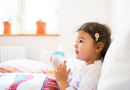 Cute little girl holding bottle with tea at home sitting on sofa in living roomの写真素材