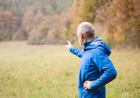 Senior runner in blue jacket resting outside in sunny autumn nature, pointing at something with his finger.の写真素材
