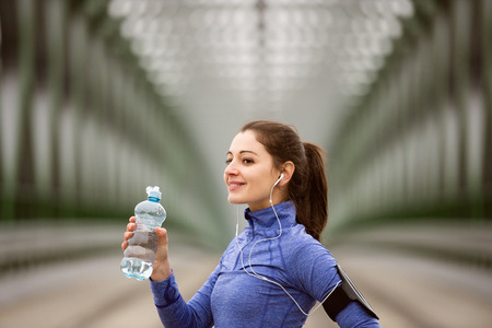 Beautiful young woman with smart phone and earphones, listening music, running in the city on green steel bridge, resting, drinking water from bottle.の写真素材