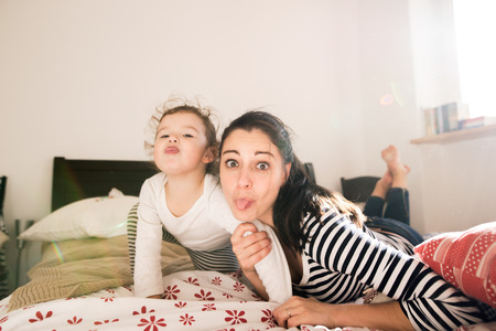 Beautiful young mother having fun with her cute little daughter on bed in her bedroom, sticking tongues outの写真素材
