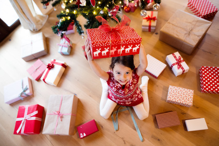 Beautiful little girl under Christmas tree sitting among presents, holding one above her head.の写真素材