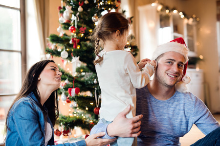 Beautiful young family with little girl decorating Christmas tree at home. Daughter giving father Christmas ornaments on his ears.の写真素材