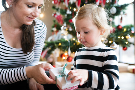 Beautiful young mother giving her cute litte daughter Christmas present at illuminated and decorated Christmas tree.の写真素材