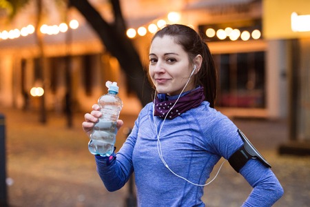 Beautiful young woman with smart phone and earphones, listening music, resting, drinking water from bottle. Running in the illuminated night town.の写真素材