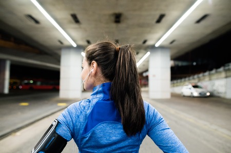 Beautiful young runner under the bridge in the town in the evening with smart phone and earphones, listening music. Using a fitness app for tracking weight loss progress, running goal or summary of her run. Resting, arms on hips. Rear view.の写真素材