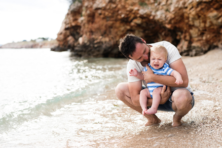Handsome young man holding his baby son at the beach enjoying time at seaside. Little boy crying.の写真素材