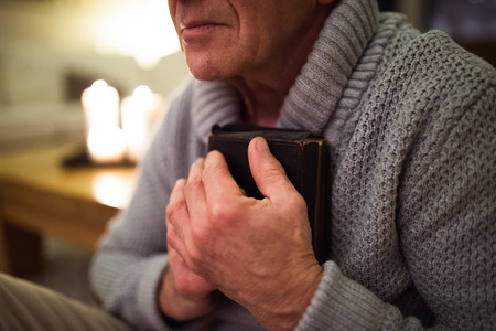 Senior man praying, holding Bible, burning candles behind himの写真素材