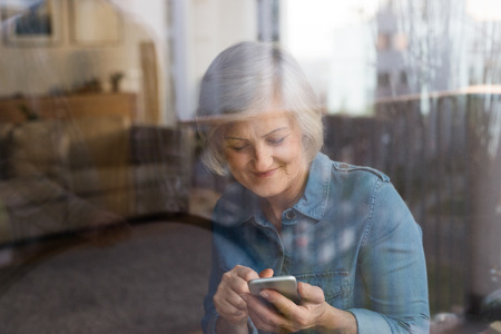 Senior woman with smartphone at home sitting at the window