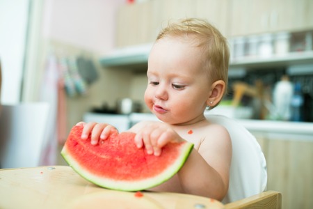 Cute little girl sitting in high chair eating watermelon