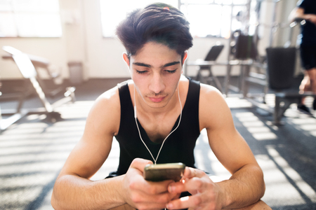 Hispanic man in gym resting, holding smart phone, listening musiの写真素材