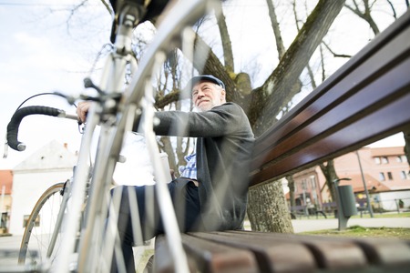 Senior man with bicycle in town, sitting on bench with coffeeの写真素材