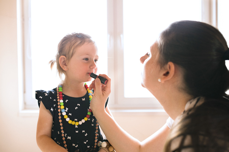 Mother painting lips of her cute little daughter with lipstick.の写真素材