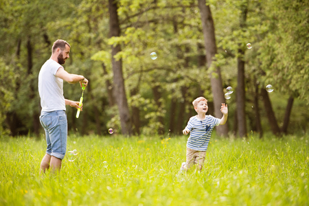 Hipster father and son blowing bubbles outdoors in park.の写真素材