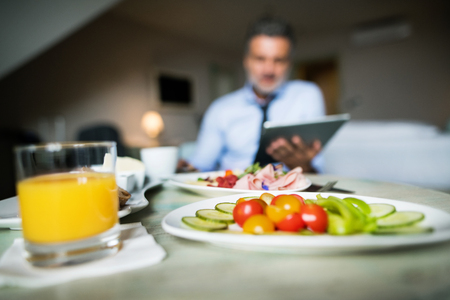 Mature, handsome businessman working on a tablet in a hotel room. A man having breakfast.の写真素材