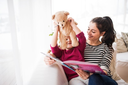 Young mother and a small girl with teddy bear at home, reading a book.の写真素材