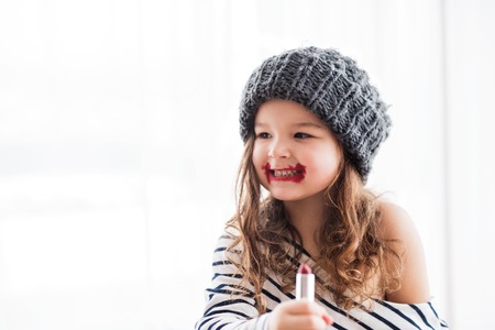 A small happy girl in striped T-shirt at home applying a lipstick.の写真素材