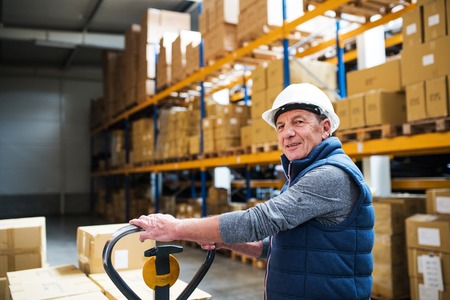 Senior male warehouse worker or a supervisor pulling a pallet truck with boxes.
