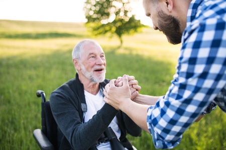An adult hipster son with senior father in wheelchair on a walk in nature at sunset.の写真素材
