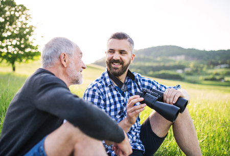 An adult hipster son with binoculars and senior father sitting on the grass in sunny nature.の写真素材