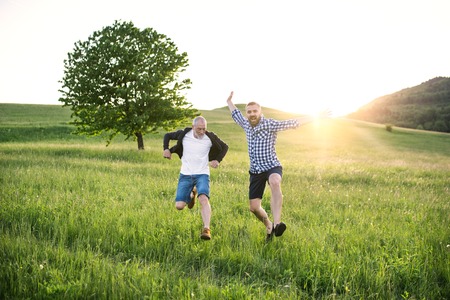 An adult hipster son and his senior father jumping in nature at sunset.の写真素材